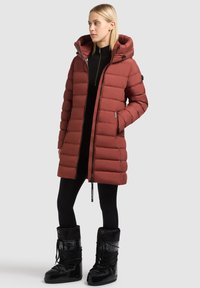 Doudoune rouge avec capuche, manches longues, deux poches latérales et fermeture éclair, associée à des bottes de neige noires et des leggings.