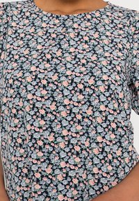 Blusa floreale navy con piccoli fiori rosa e blu, scollo rotondo, maniche corte e dettaglio arricciato; il tessuto sembra leggero e strutturato.