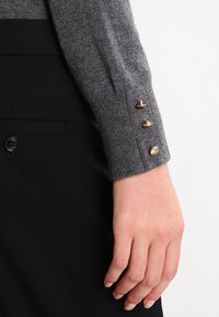 Manche de pull en maille grise avec une finition texturée. Quatre boutons dorés ornent le poignet, associés à un pantalon noir avec un détail de bouton.