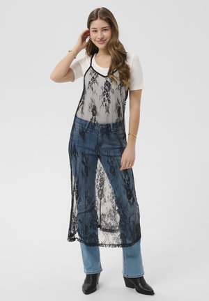 Tânără purtând tricou alb cu mâneci scurte, suprapus cu rochie lungă neagră din dantelă, blugi albaștri și cizme negre până la gleznă, stând cu mâna atingându-și părul.