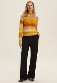 Maglione lavorato a maglia multicolore con strisce gialle, rosa e arancioni, maniche lunghe e una finitura testurizzata, abbinato a pantaloni neri e scarpe flat.
