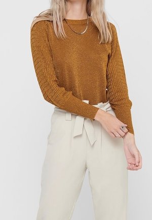 Femme portant un pull à manches longues métallique couleur rouille et un pantalon taille haute crème style paperbag avec ceinture nouée, ajustant sa manche avec la main.