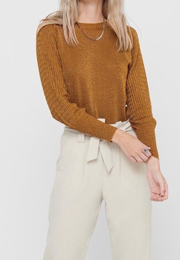 Femme portant un pull à manches longues métallique couleur rouille et un pantalon taille haute crème style paperbag avec ceinture nouée, ajustant sa manche avec la main.