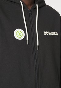 Schwarze Zip-Hoodie mit weißen Kordeln, auf der "DCSHOECO" in Weiß gestickt ist und einem grünen runden Etikett, das umweltfreundliches Material anzeigt.