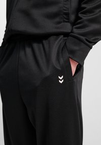 Svarta sportbyxor i slät tyg, med en sidficka och en distinkt ljusfärgad chevron-design på låret.