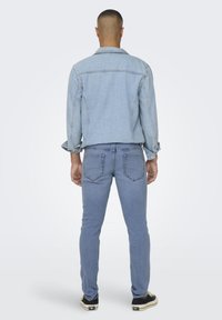 Lys blå denimjakke med standard krage, kombinert med lys blå jeans. Begge plaggene har sydd detaljer. Skoene er svarte med hvite aksenter.