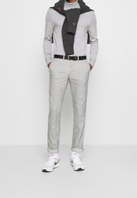 Man i ljusgrå polotröja, grå byxor med svart bälte, mörkgrå sweater över axlarna, vita Nike-sneakers.