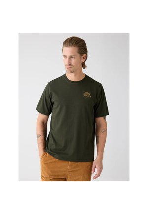 Man met tatoeages draagt een donkergroen T-shirt met een kleine gele afbeelding op de borst en mosterdkleurige corduroy broek, kijkt iets naar links.