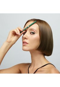 Crayon eye-liner vert tenu contre le front, coupe de cheveux courte et droite châtain, fond neutre, mise au point sur le visage et le maquillage.