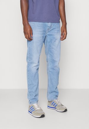 Person, die hellblaue Jeans, beige-blaue Sneakers und ein lila Hemd trägt, steht vor einem schlichten hellen Hintergrund.