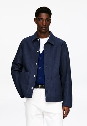 Veste en denim bleu marine avec une fermeture à boutons, deux poches avant et un col classique. Portée par-dessus un pull bleu et un pantalon blanc.