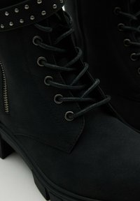 Acercamiento de botas de tobillo negras con cordones, suelas gruesas, ojales de metal, cremallera y correa con tachuelas en un material mate liso.