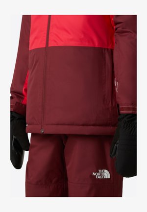 Zimska jakna boje tamnocrvene i crvene s crnim rukavicama. Sadrži patentne zatvarače, elastične manžete i logotip na hlačama. Vodootporni materijal.