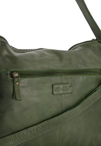 Groene leren tas met een gladde textuur, voorzien van een voorzak met rits en een logo-opdruk. Verstelbare schouderband in bijpassend groen.