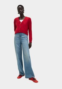 Cardigan rossa in lana con scollo a V e bottoni dorati, abbinato a jeans a vita alta blu chiaro con gamba larga e ballerine rosse con ritagli.