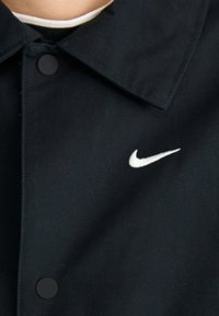 Crna pamučna jakna s šiljastim ovratnikom, koja ima bijeli Nike logo na gornjoj lijevoj strani i crne kopče na dugmad.