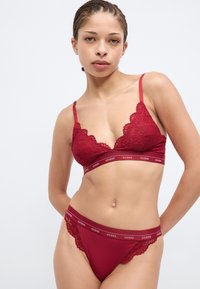 Red lace bralette and matching thong with scalloped edges and elastic band featuring "GUESS" logo. Soft, textured fabric with adjustable straps.