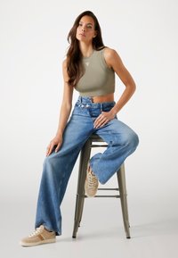Olivgrön ärmlös crop top, ljusblå vida jeans och beige sneakers, sittande på en grå metallpall, minimal bakgrund.