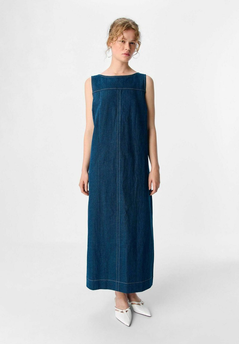 Dolga denim obleka v temno modri barvi s ravnim krojem, širokim ovratnikom in podrobnostmi na stranskih šivih. Oblečena z belimi čevlji z izrazitim nosom.