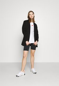 Blazer nero, t-shirt bianca con grafica di labbra rosse, pantaloni shorts grigi per bici, sneaker bianche con accenti arancioni, in piedi su uno sfondo neutro.