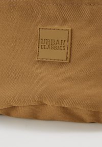 Brun tekstilpose med en struktureret overflade, der har et firkantet, præget "URBAN CLASSICS" logo i en læderlap i tan.