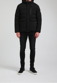 Czarna kurtka puchowa z kapturem, zapinaną na zamek kieszenią na piersi, dwoma przednimi kieszeniami oraz pikowanym wzorem. Noszona z dopasowanymi czarnymi dżinsami i czarnymi trampkami.