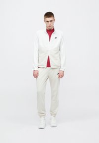 Chaqueta blanca con cremallera y diseño triangular, camiseta roja debajo, pantalones beige y zapatillas blancas. Presenta el logo de Nike en el pecho.