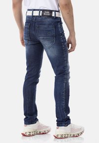 Jean en denim bleu foncé à coupe ajustée, avec un design de poche arrière distinctif et une ceinture blanche avec des lettres noires.