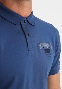 Polo en coton bleu marine avec col, doté d'une patte à deux boutons, texte brodé "SUNSET CAL" et détails de couture contrastants.