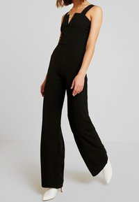 Combinaison noire avec encolure structurée et jambes larges. Fabriquée à partir d'un tissu lisse. Portée avec des talons blancs pointus et des bijoux minimalistes.