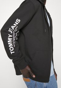 Haut de hoodie zippé noir en mélange de coton, avec le texte blanc "TOMMY JEANS" le long de la manche gauche et un détail de poche kangourou à l'avant.