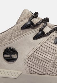 Zapatillas beige que cuentan con una parte superior de ante texturizado, ojales negros y un logo con un emblema de árbol. Los cordones son oscuros con un patrón y la suela es de color blanco roto.