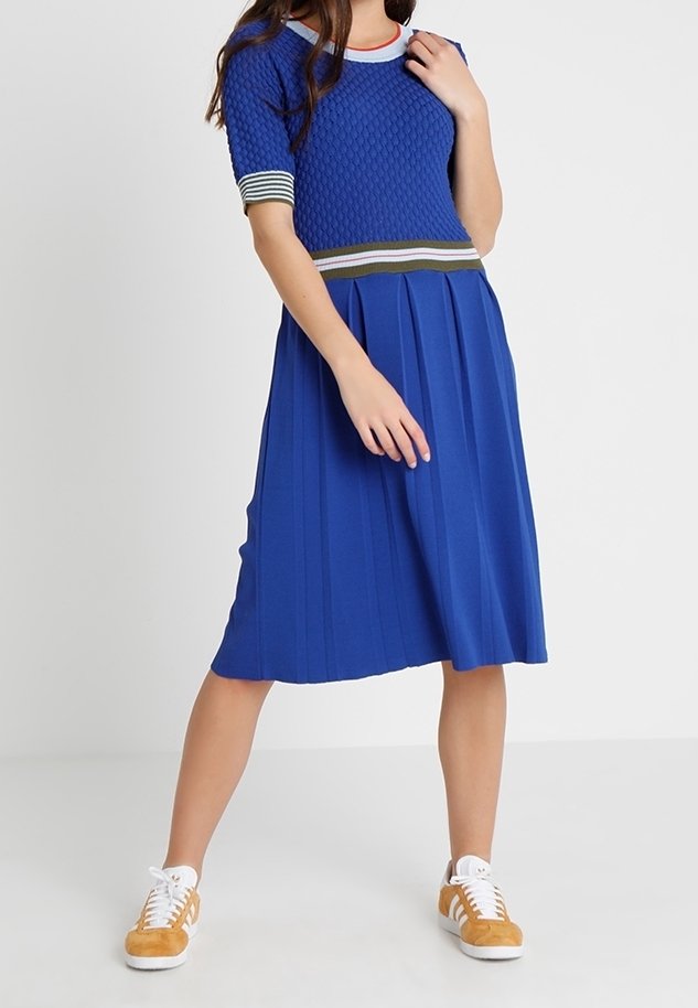 Femme portant un haut en tricot bleu texturé et une jupe plissée avec un bord rayé, associée à des baskets jaune moutarde et blanches, debout, les jambes écartées.