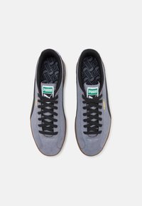 Gray suede sneakers with black laces, gum rubber soles, and a green logo patch. Features a textured inner sole with brand pattern.