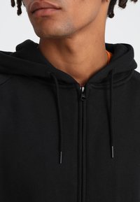 Sudadera negra con cremallera y un tejido suave y texturizado, que presenta una capucha con cordón y un forro interior naranja en contraste.