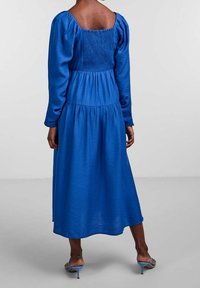 Vestido azul de manga larga con espalda cuadrada, corpiño fruncido y falda en capas, hecho de tela texturizada. Acabado con sandalias de tacón alto.