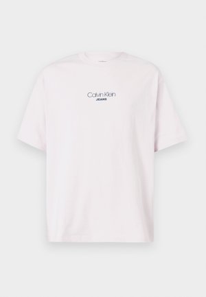 T-shirt in cotone rosa chiaro a maniche corte con logo testuale "Calvin Klein JEANS" centrato sul petto su uno sfondo semplice.