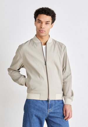Veste bomber beige clair avec fermeture éclair à l'avant, poignets à côtes et ourlet. Associée à un jean en denim bleu, sur un fond neutre.