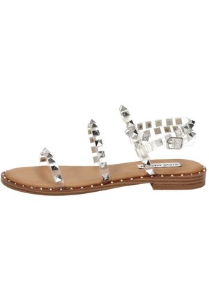 Przezroczyste sandały na płaskiej podeszwie z metalicznymi srebrnymi ćwiekami, brązową wkładką i zapięciem na klamrę. Teksturowane akcenty na paskach i krawędzi.