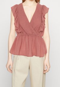 Rynkad, V-ringad topp i dammig ros med lätt, texturerad tyg; elastisk midja som skapar en peplumform, tillsammans med beige byxor.