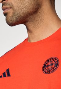 Piros pamut póló kerek nyakkivágással és rövid ujjakkal. Navy FC Bayern München logóval és három vízszintes navy csíkkal a bal ujján.