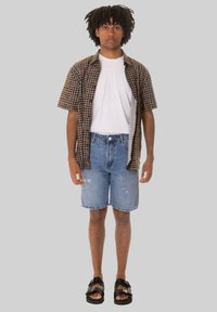 Camicetta a maniche corte a scacchi con un motivo marrone e nero, t-shirt bianca, pantaloncini in denim e sandali neri con suola antiscivolo.
