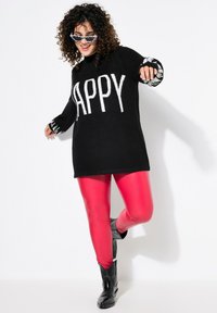 Maglione nero lavorato a maglia con la scritta "HAPPY" in bianco, abbinato a leggings rossi lucidi e stivali da cowboy neri, con dettagli a strisce sulle maniche.