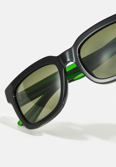 Occhiali da sole neri oversize con lenti verde scuro e dettagli verdi sulle tempie. Finitura liscia e lucida con design del telaio angolare.