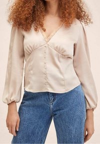 Femme aux cheveux roux bouclés portant un chemisier en satin crème avec des boutons et des bordures en dentelle, associé à un jean bleu taille haute.