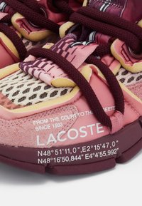 Close-up af pink og bordeaux Lacoste sneaker med mesh, ruskindsdetaljer, bordeaux snørebånd og trykt tekst inklusive koordinater på sålen.