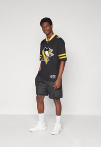 Fekete hoki mez sárga csíkokkal és egy pingvin logóval, fekete hálós rövidnadrággal és fehér sportcipőkkel párosítva, egy szürke háttér előtt állva.