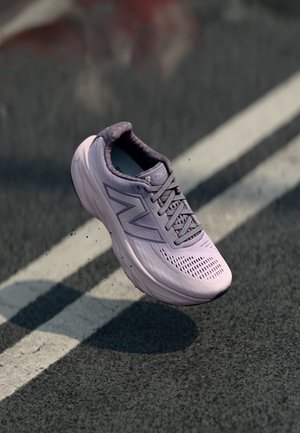 Enkele lavendelkleurige New Balance hardloopschoen zwevend boven een asfaltweg met witte lijnmarkeringen.