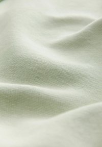 Világoszöld szövet sima textúrával, amelyen finom szőtt minta látható. Az anyag rugalmasnak és könnyűnek tűnik.