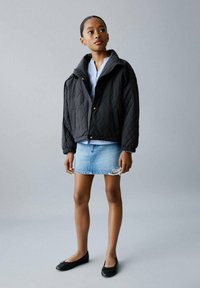 Veste matelassée noire avec un col montant, superposée à une chemise à rayures bleues, associée à une jupe en denim clair et des ballerines noires. Design simple.